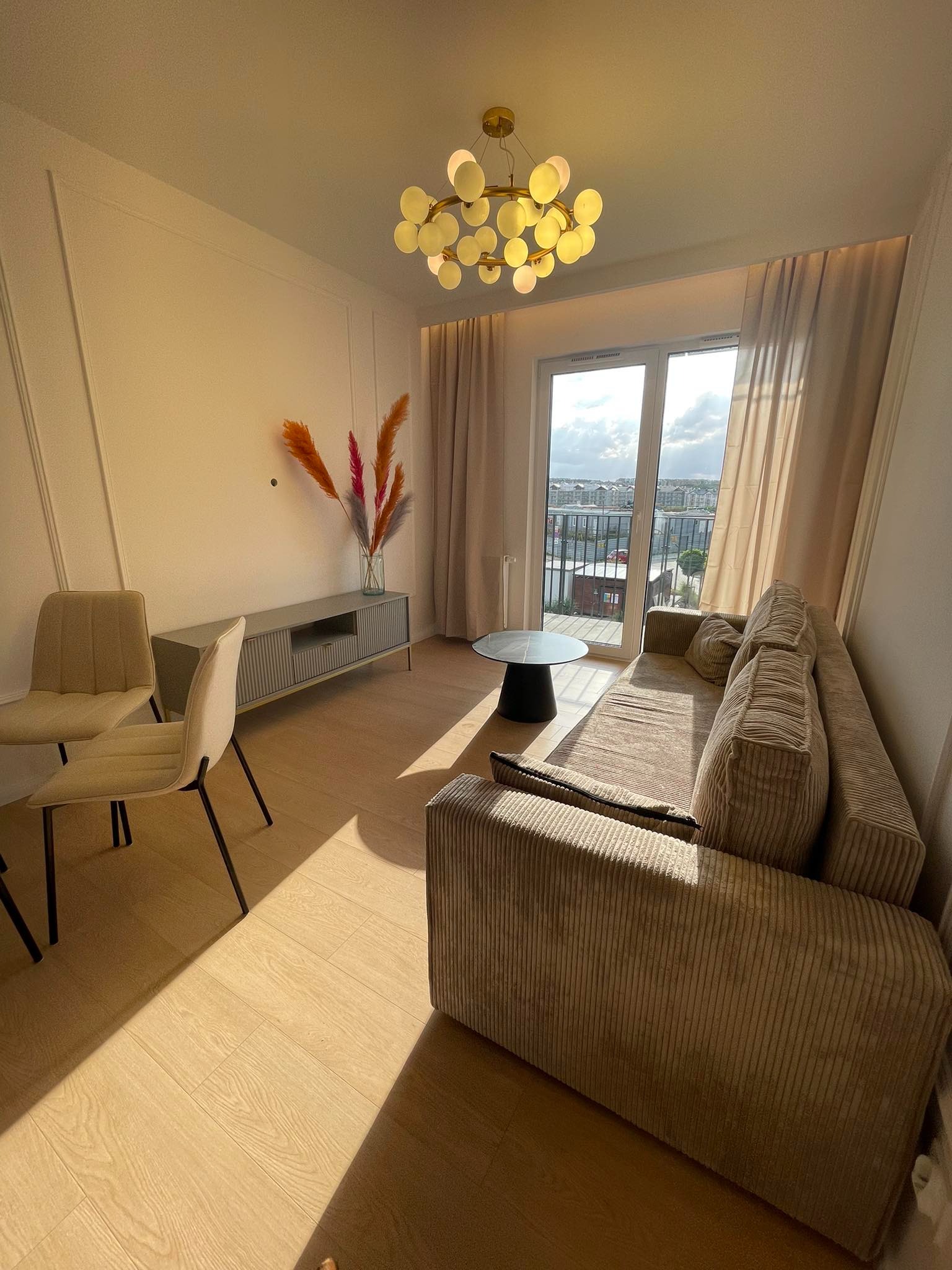 Jasne wnętrze salonu z beżową sofą, stolikiem kawowym i krzesłami. Widok na miasto przez okno balkonowe. Dekoracyjne listwy na ścianie i nowoczesna lampa.