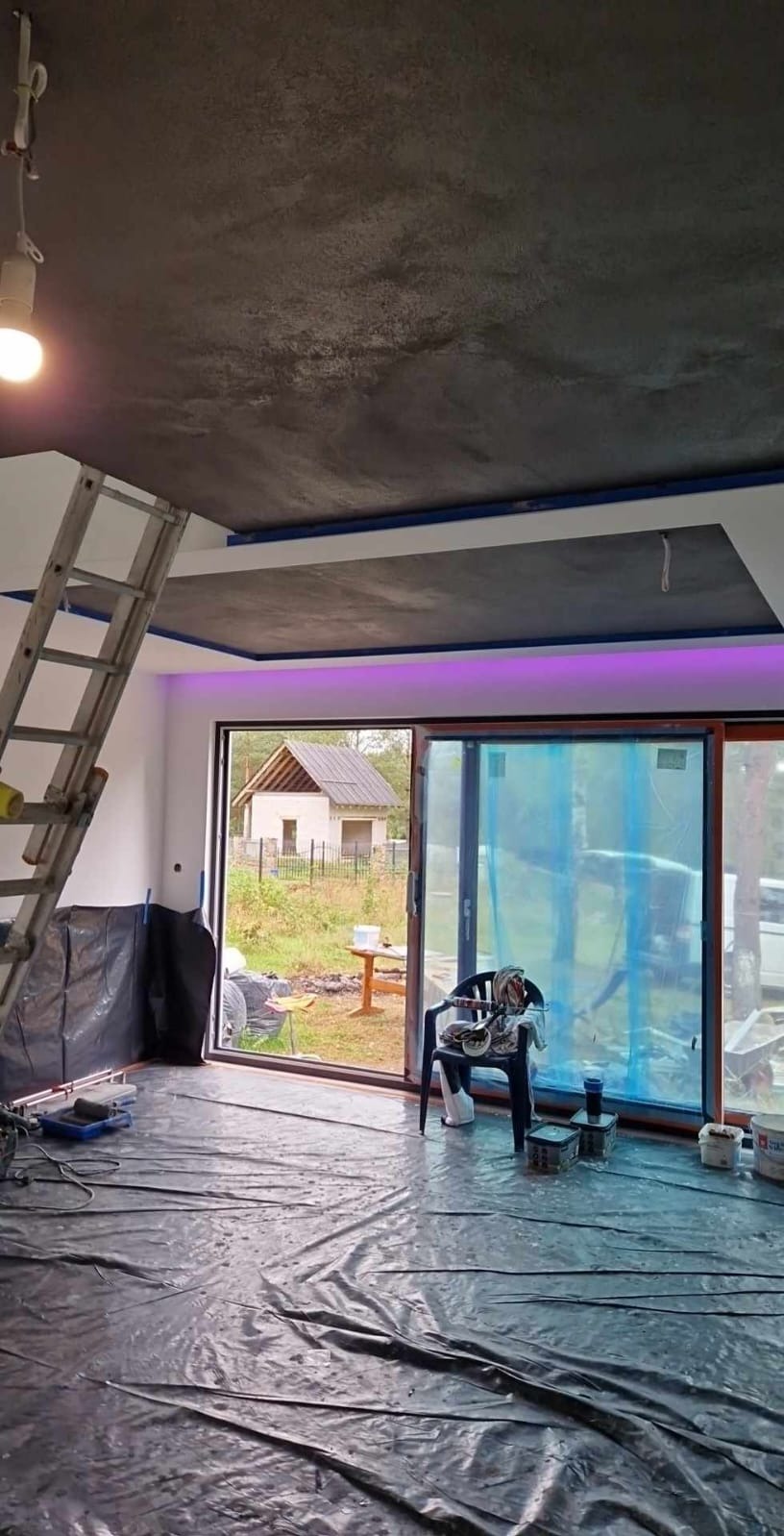 Pokryty folią pokój w trakcie prac wykończeniowych. Widoczna drabina, narzędzia malarskie i sufit z efektem dekoracyjnym. W tle widok na zewnątrz przez duże okno.