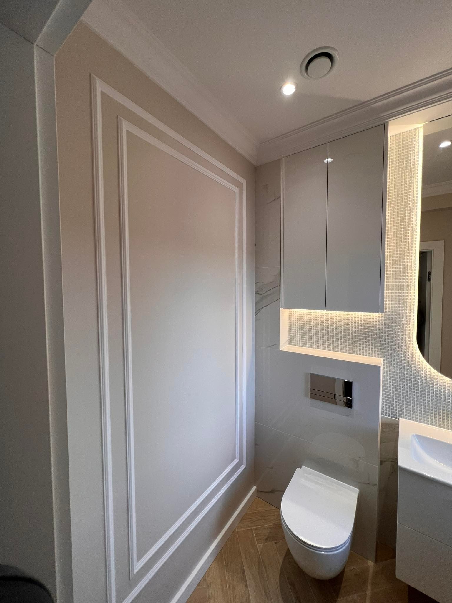 Elegancka łazienka w odcieniach bieli i beżu. Dekoracyjne listwy ścienne, podświetlane lustro i szafka nad toaletą. Parkiet ułożony w jodełkę.