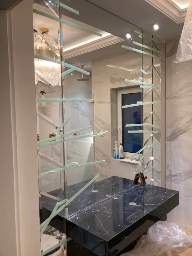 Nowoczesna łazienka z lustrem w dekoracyjnych taflach, zabezpieczonych taśmą. Czarny blat z marmurowym wzorem, białe marmurowe płytki na ścianach. Widoczny fragment żyrandola.