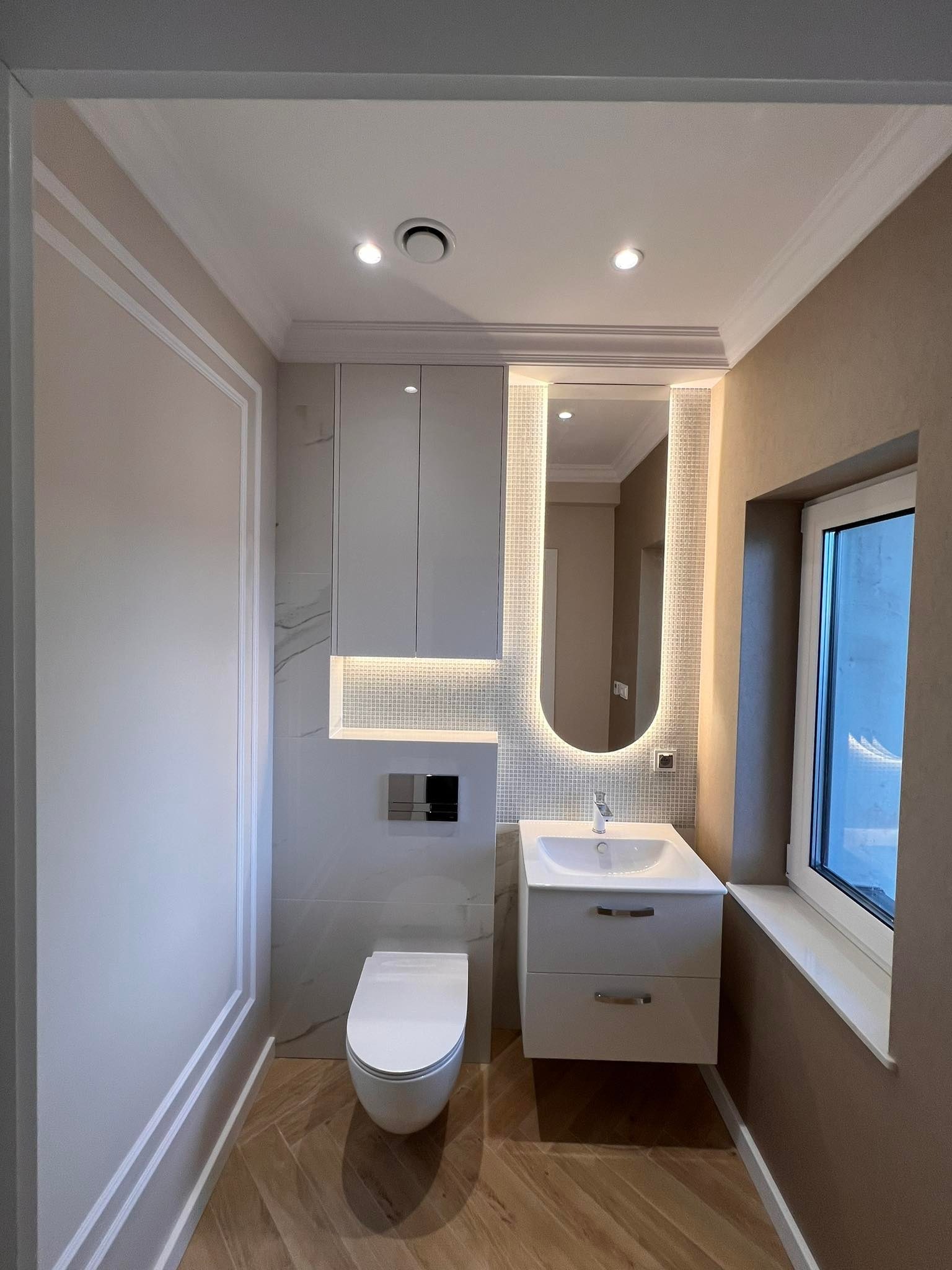 Elegancka łazienka w Słupsku po remoncie: biała armatura, podświetlane lustro, płytki imitujące marmur i drewnopodobna podłoga ułożona w jodełkę.