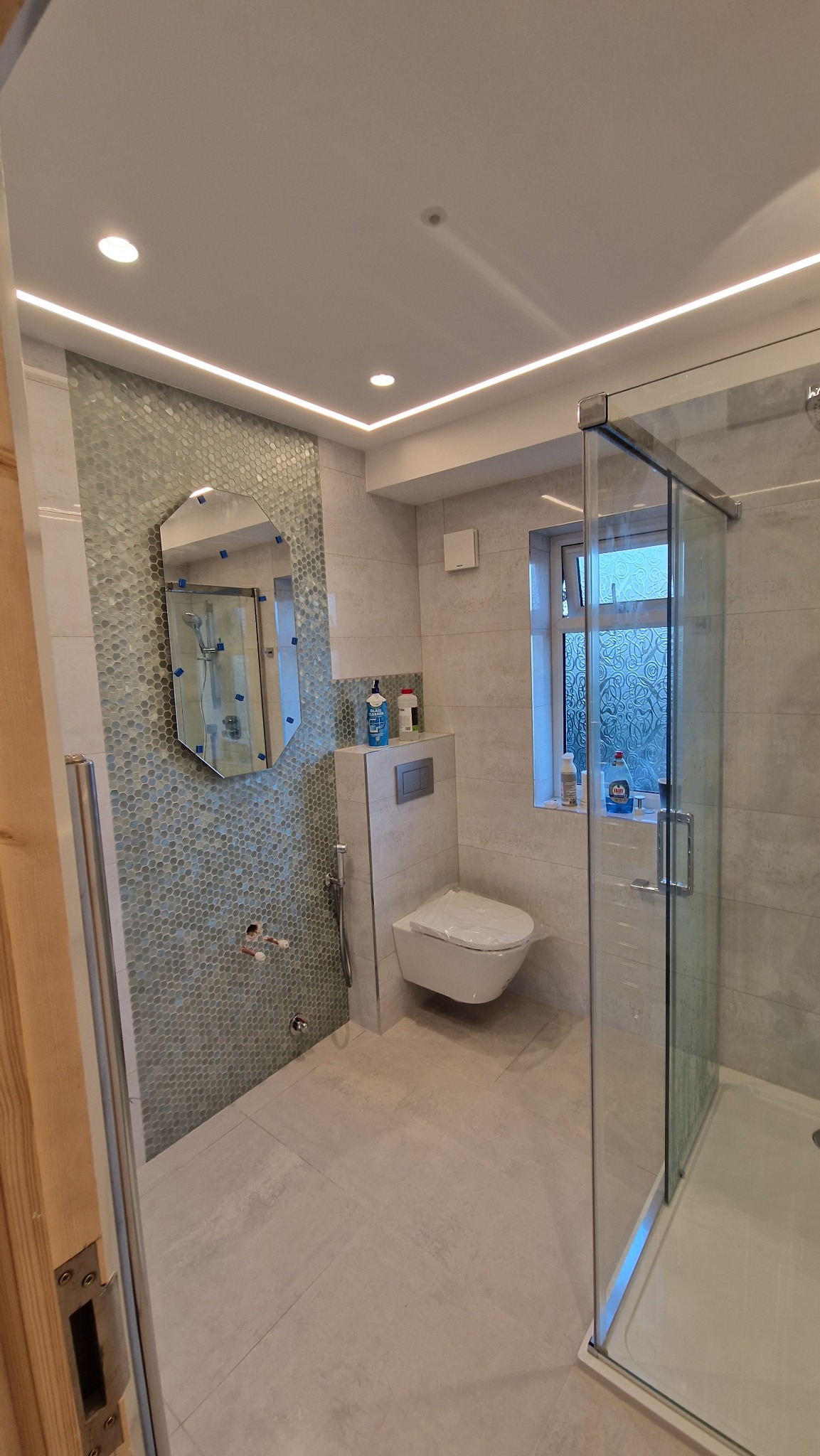 Nowoczesna łazienka z kabiną prysznicową, mozaiką na ścianie i podświetleniem LED. Umywalka i toaleta w minimalistycznym stylu. Lustro w ozdobnej ramie.