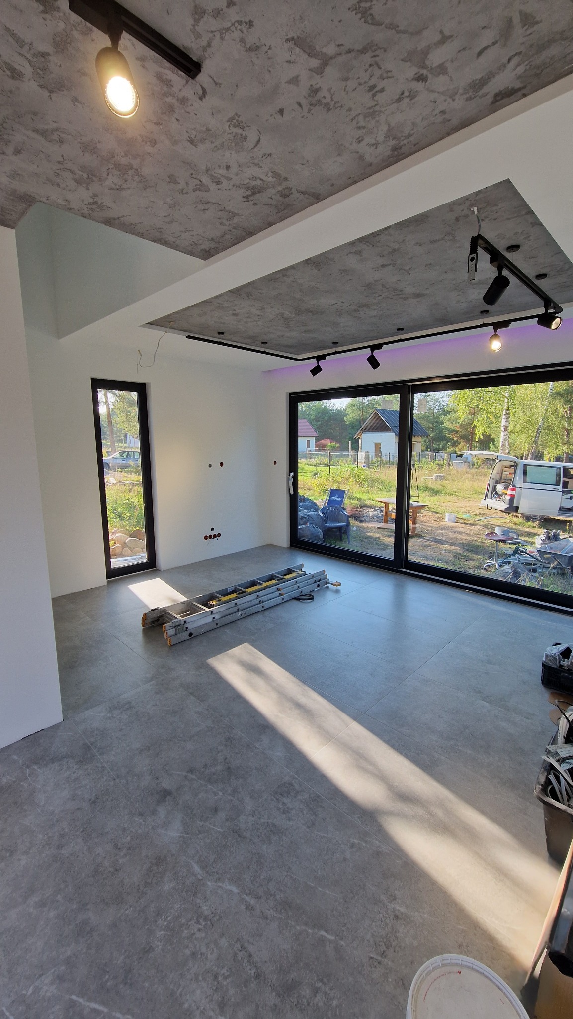 Surowe wnętrze z szarą podłogą i sufitem o teksturze betonu. Rozłożona drabina i widoczne punkty elektryczne sugerują etap prac wykończeniowych.