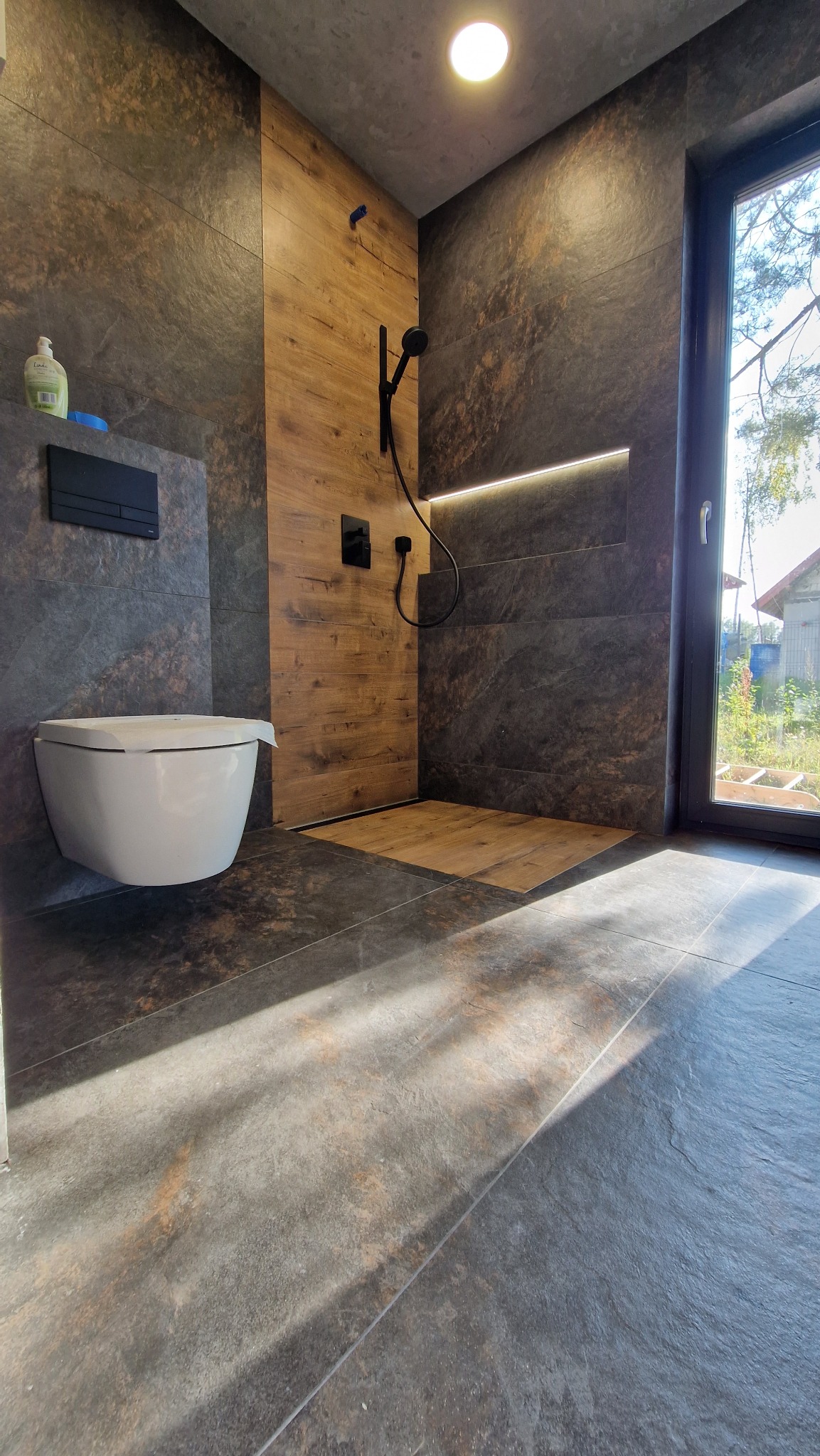 Nowoczesna łazienka z kabiną typu walk-in, wykończona płytkami imitującymi kamień i drewno. Uwagę zwraca podświetlenie LED w niszy i czarna armatura.