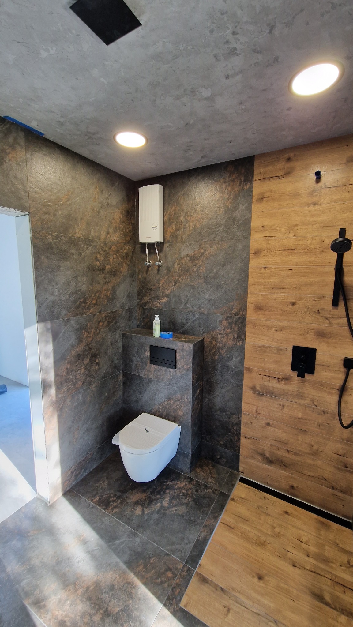 Nowoczesna łazienka z płytkami imitującymi kamień i drewno. Biała toaleta podwieszana, czarna armatura prysznicowa. Oświetlenie punktowe w suficie.