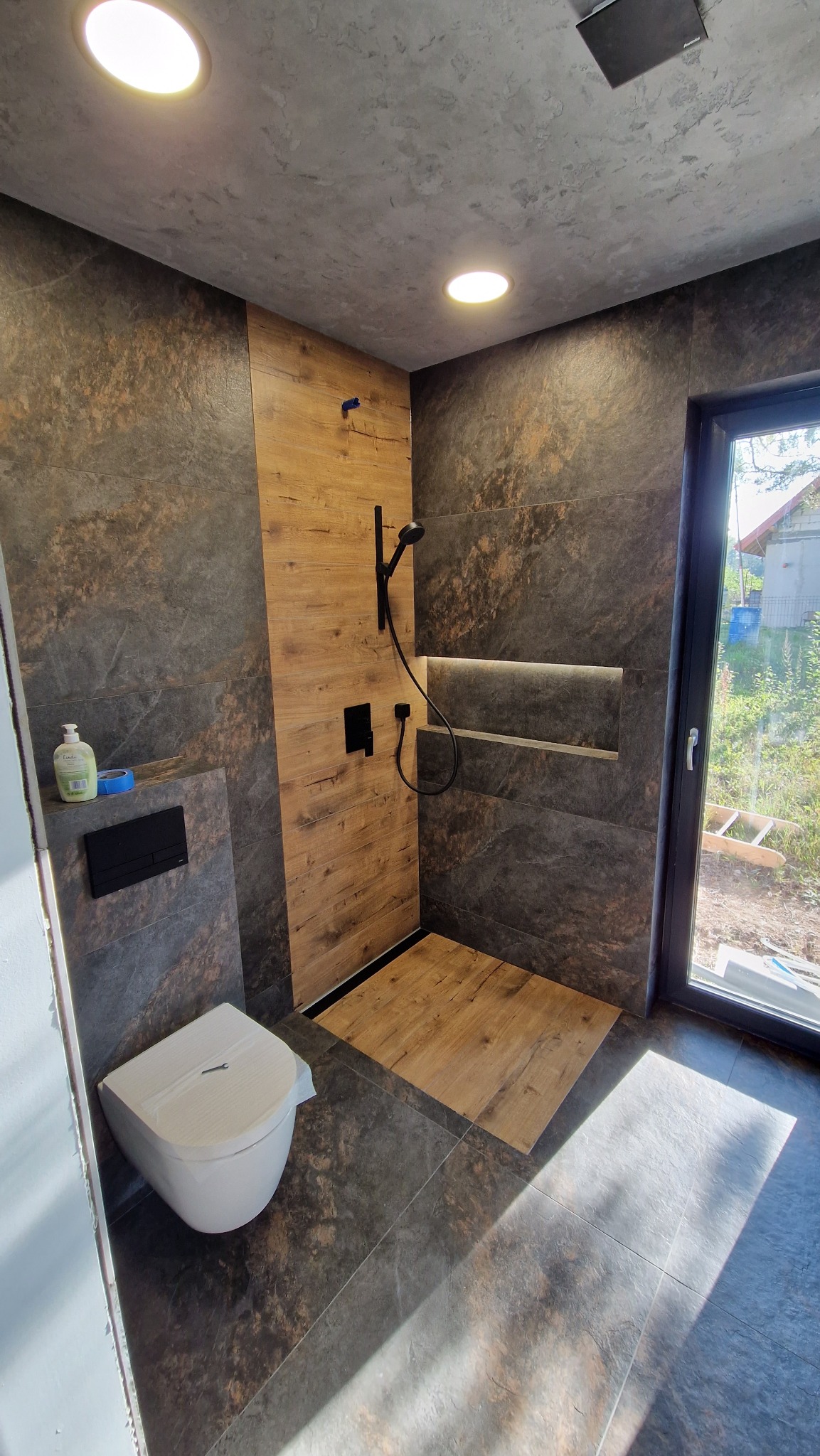 Nowoczesna łazienka z kabiną typu walk-in, wykończona płytkami imitującymi drewno i kamień. Uwagę zwraca czarna armatura i oświetlenie LED w półce.