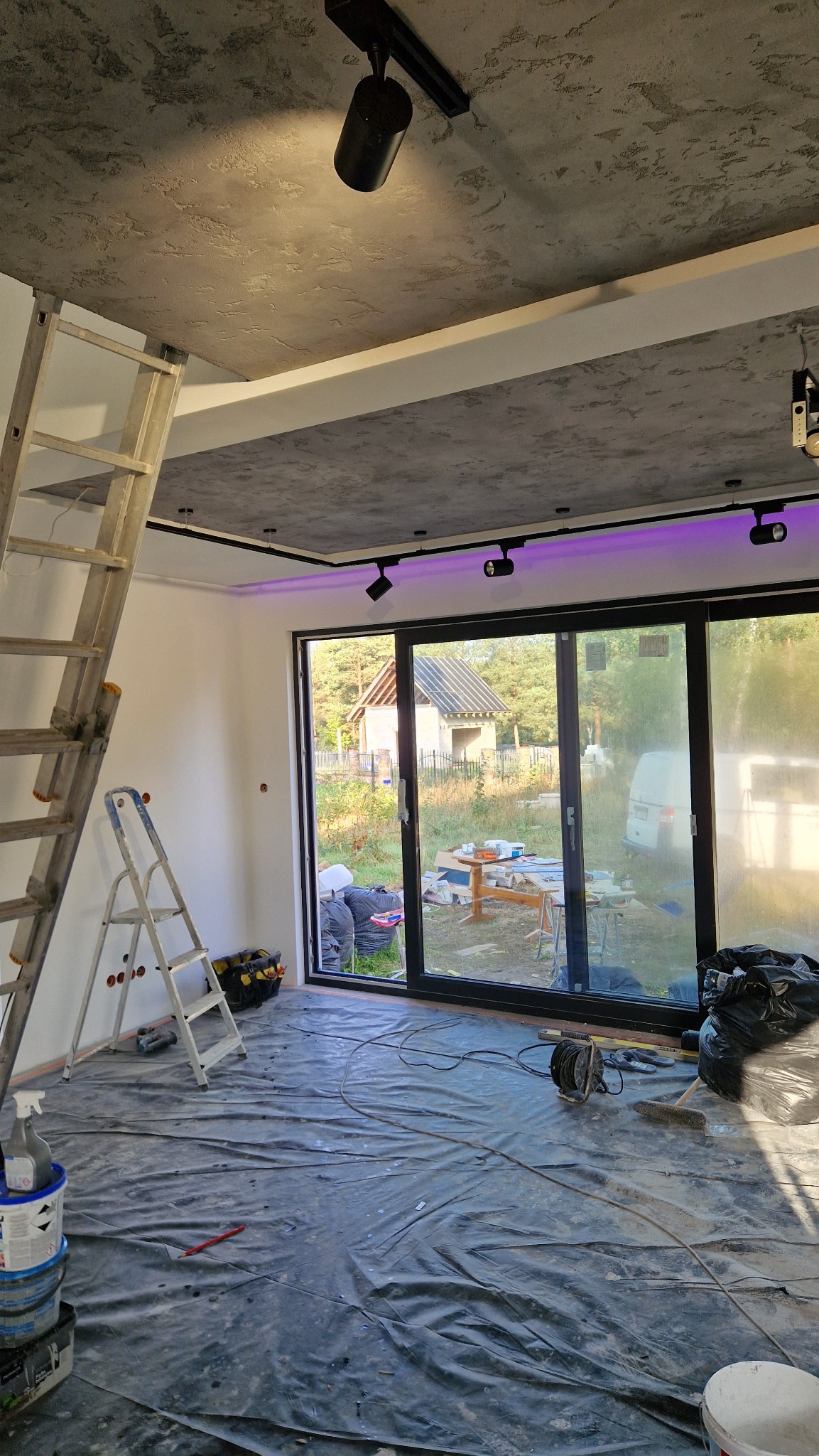 Pokój w trakcie prac wykończeniowych: zabezpieczona podłoga folią, drabiny, widoczne narzędzia i sufit z oświetleniem punktowym. Widok na zewnątrz przez duże okno.