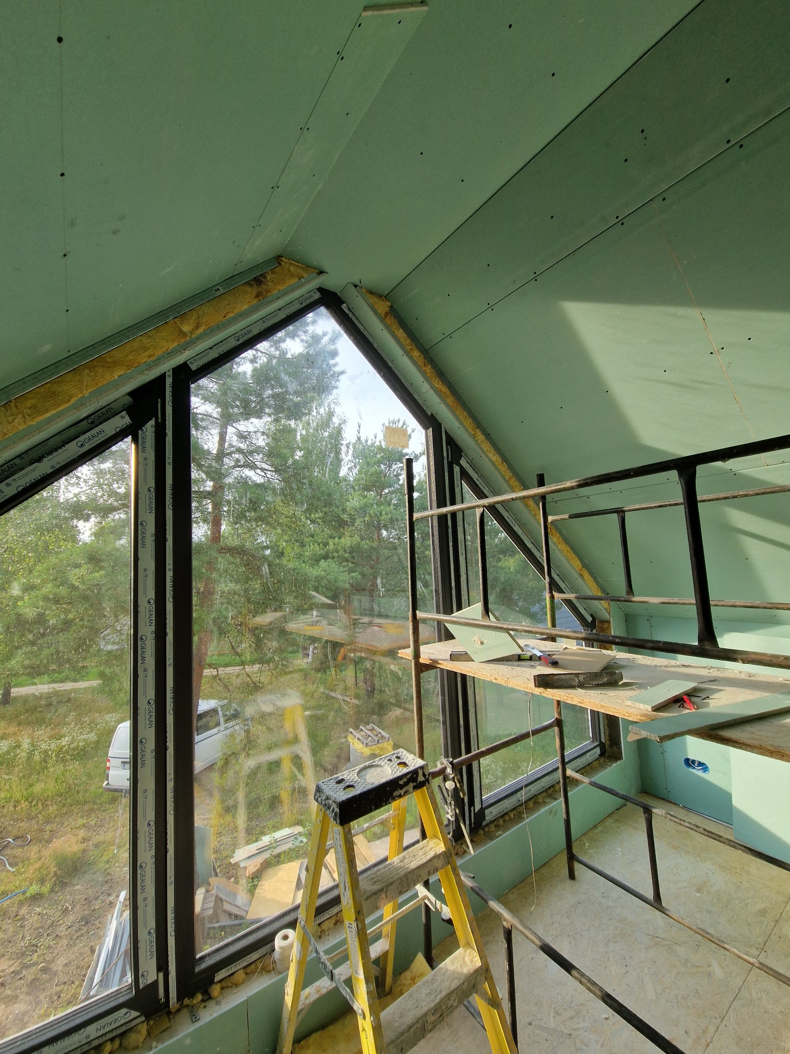 Montaż zielonej płyty karton-gips na poddaszu z oknem, widoczne rusztowanie i drabina. Prace wykończeniowe w nowym domu z widokiem na las.