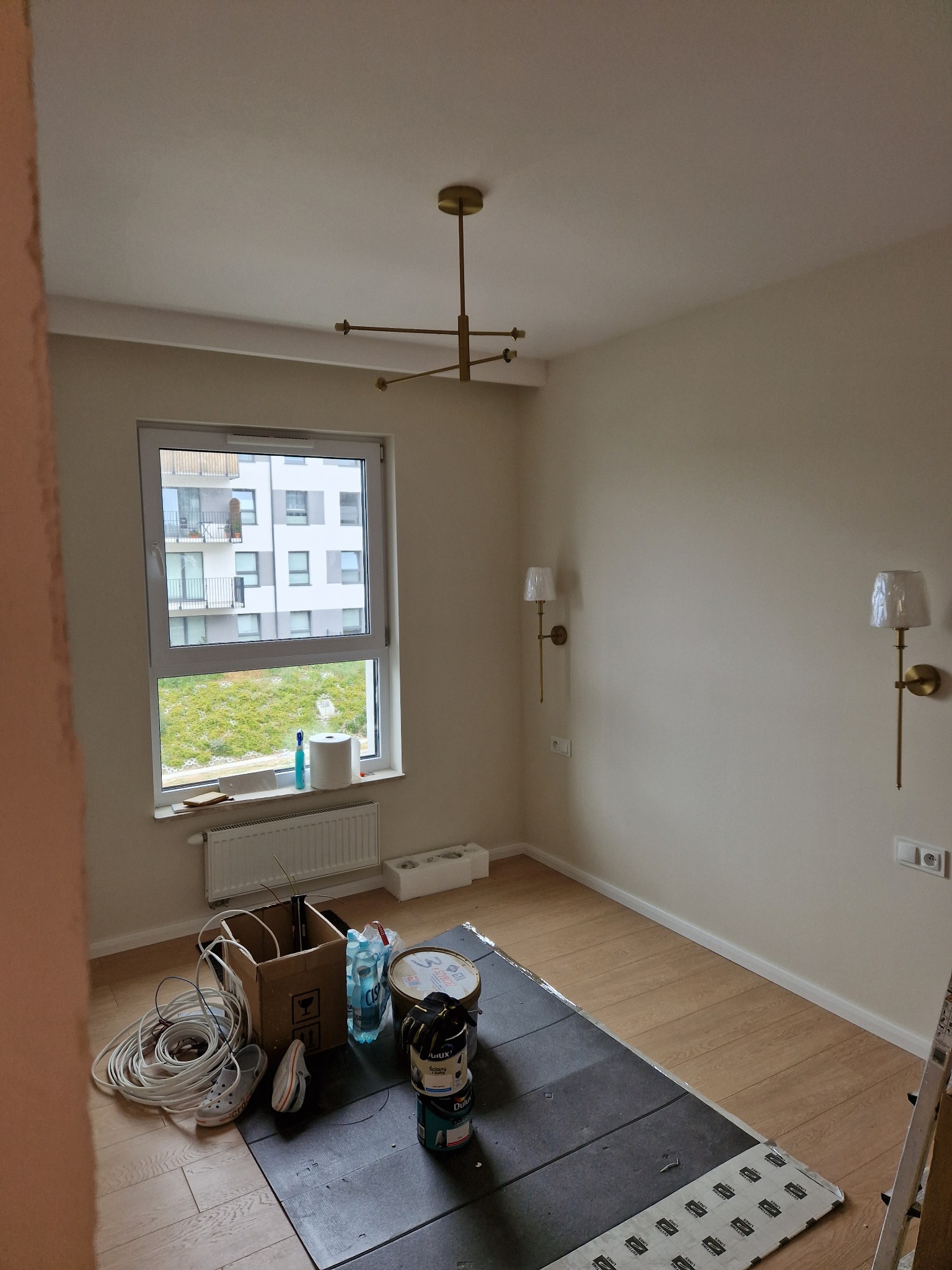 Jasne wnętrze w trakcie prac: położone panele podłogowe, puszki z farbą, lampy ścienne w folii, okno z widokiem na budynek. Etap przed ostatecznym wykończeniem.