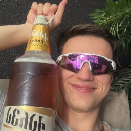 Patryk Dobrakowski - Uśmiechnięty mężczyzna w okularach trzyma dużą butelkę piwa Karva, z odbiciem w okularach. W tle czarna ściana i roślina doniczkowa.