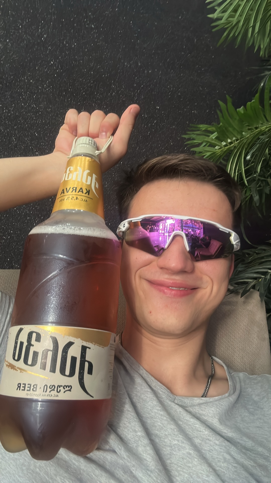 Uśmiechnięty mężczyzna w okularach trzyma dużą butelkę piwa Karva, z odbiciem w okularach. W tle czarna ściana i roślina doniczkowa.