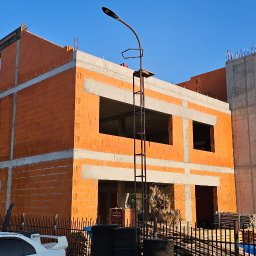 AMJ Michał Jezierski - Surowy stan dwukondygnacyjnego domu z czerwonej cegły i betonu, widoczny z zewnątrz, z elementami zbrojenia na dachu. W tle błękitne niebo i drzewa.