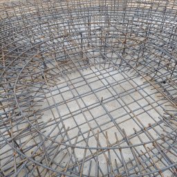 AMJ Michał Jezierski - Zbrojenie okrągłego fundamentu z prętów żebrowanych, ułożonych w siatkę. Widoczne złączenia drutem wiązałkowym. Surowy beton w tle. Detal budowy.