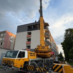 AMJ Michał Jezierski - Żółto-biały dźwig samochodowy firmy Terlik na tle bloku mieszkalnego z balkonami. Na dachu bloku widoczna sylwetka osoby. Błękitne niebo z chmurami w tle.