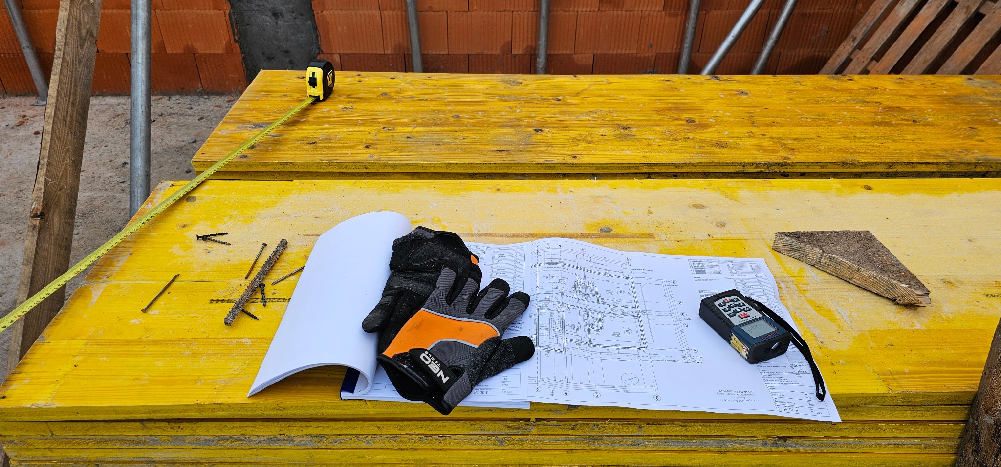 Żółte deski szalunkowe z planem budowlanym, rękawicami roboczymi, dalmierzem laserowym i miarką zwijaną. Widoczne gwoździe i wkręty. Ujęcie z budowy.