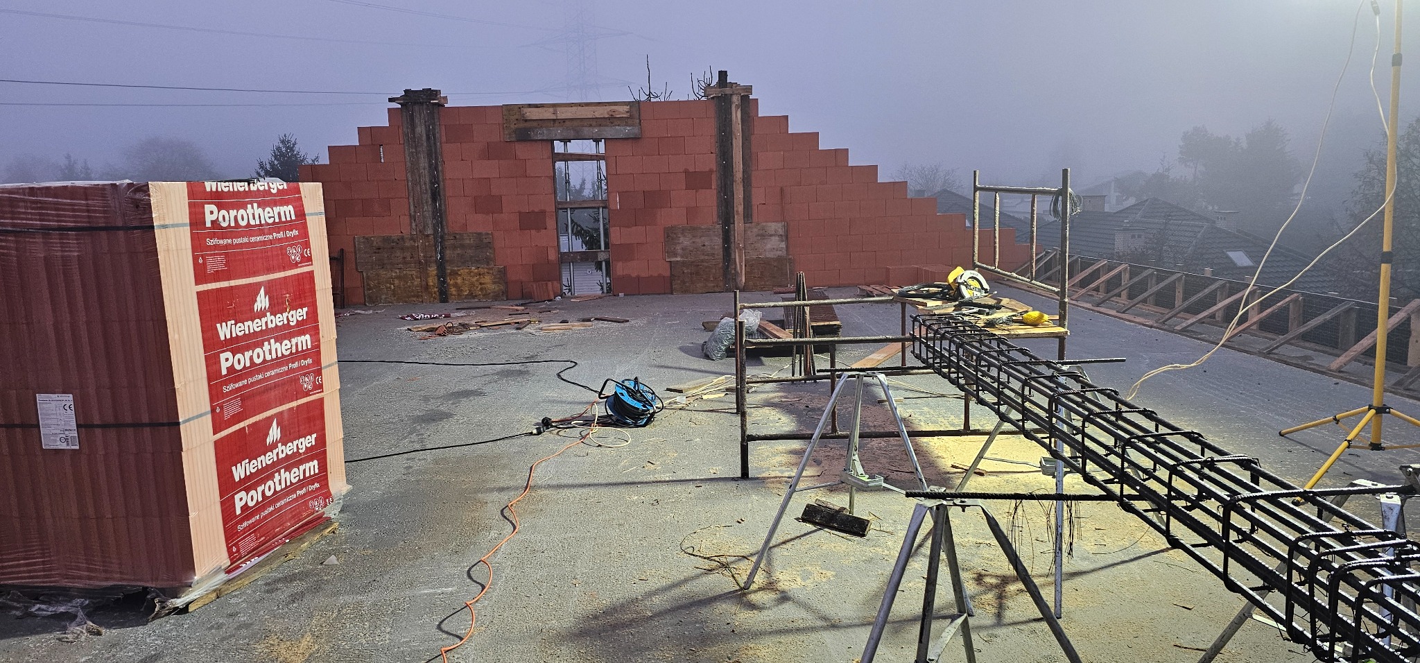 Budowa domu w trakcie realizacji: mur z pustaków Porotherm, zbrojenie na rusztowaniu, narzędzia budowlane, mgła w tle. Widok z góry na plac budowy.