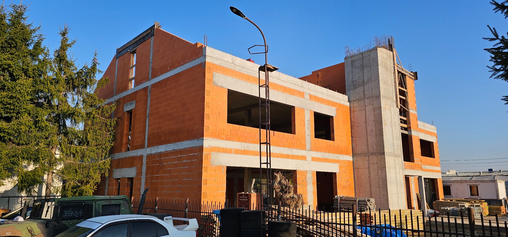 Surowy stan dwukondygnacyjnego domu z czerwonej cegły i betonu, widoczny z zewnątrz, z elementami zbrojenia na dachu. W tle błękitne niebo i drzewa.