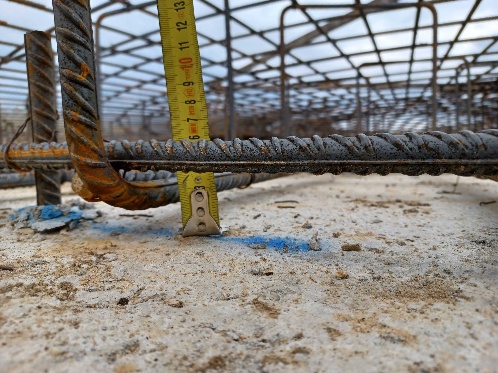 Pomiar odległości prętów zbrojeniowych za pomocą miarki na betonowej powierzchni. Widoczna siatka zbrojeniowa w tle. Bliski plan, perspektywa z dołu.