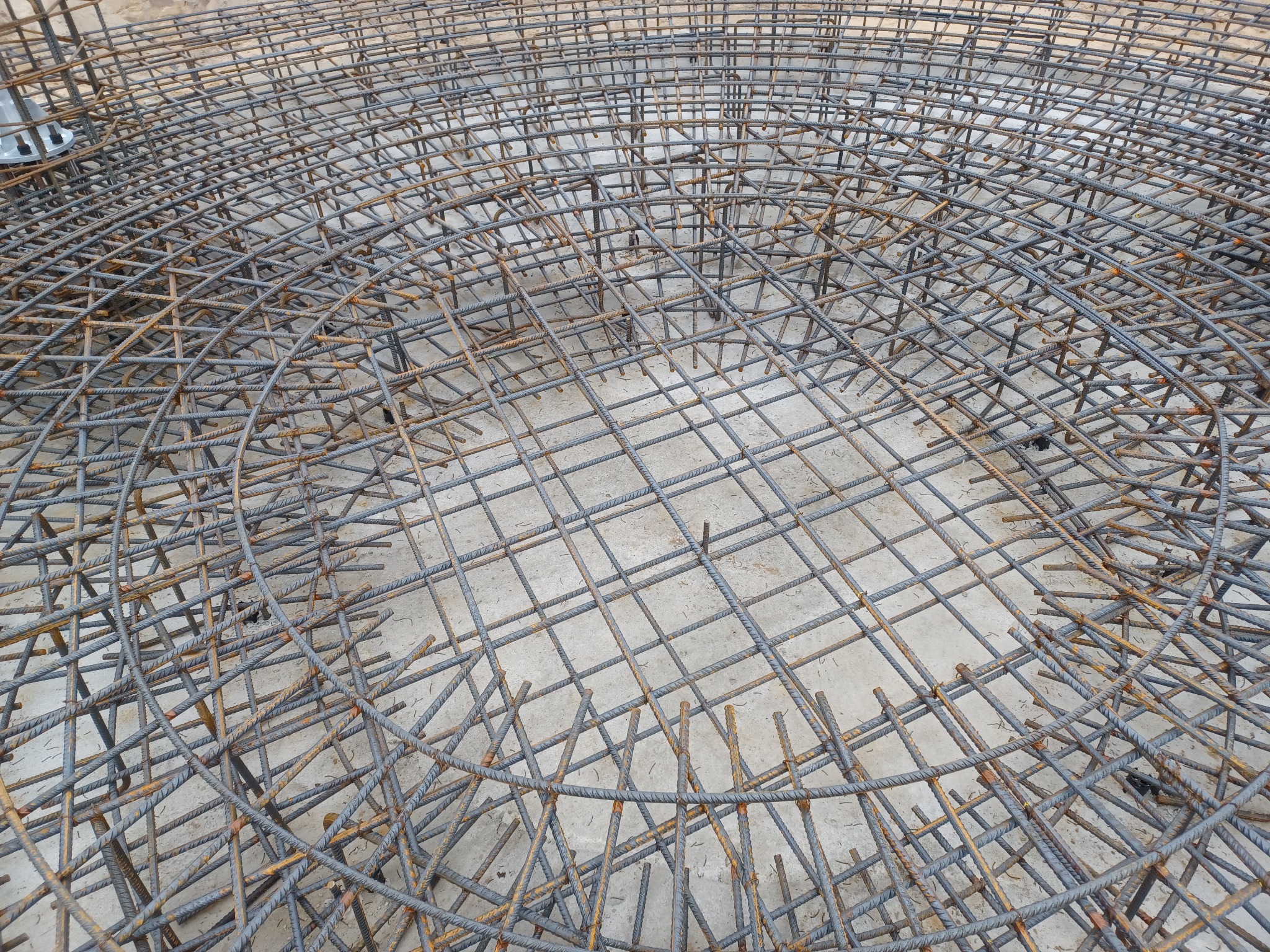 Zbrojenie okrągłego fundamentu z prętów żebrowanych, ułożonych w siatkę. Widoczne złączenia drutem wiązałkowym. Surowy beton w tle. Detal budowy.