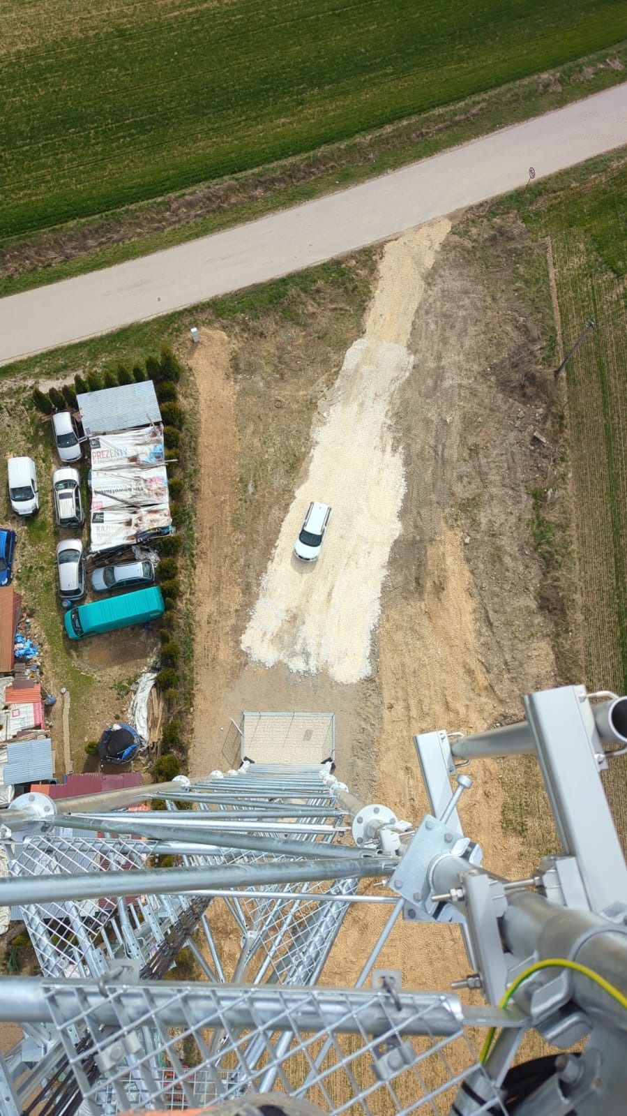 Widok z góry na plac budowy z metalowej konstrukcji wieży. Biały samochód na żwirowej drodze, budynki i pole w tle. Nietypowa perspektywa.
