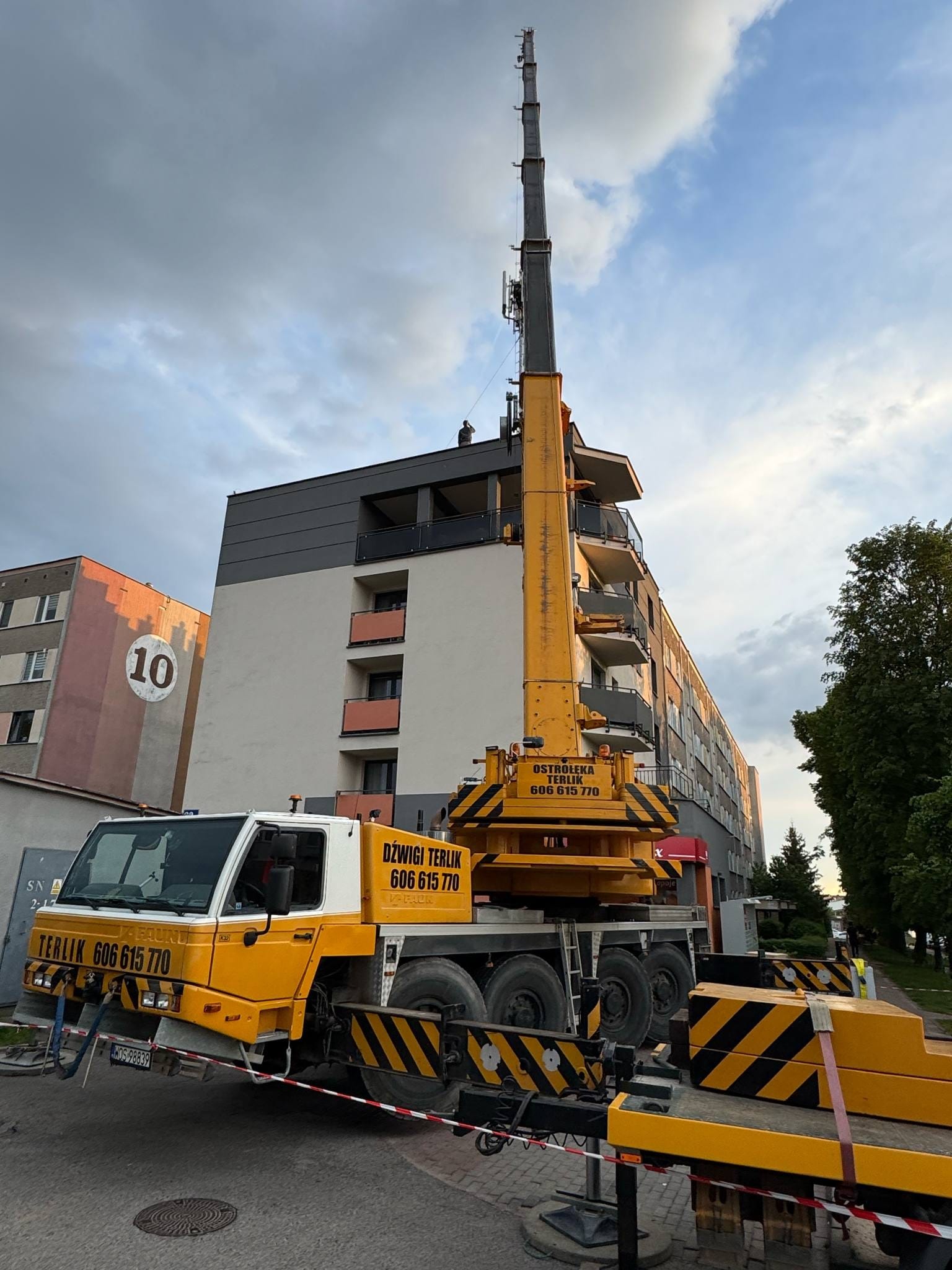 Żółto-biały dźwig samochodowy firmy Terlik na tle bloku mieszkalnego z balkonami. Na dachu bloku widoczna sylwetka osoby. Błękitne niebo z chmurami w tle.