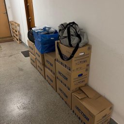 Clearbox pl - Stos kartonowych pudeł Action, torba podróżna i niebieska torba Ikea w korytarzu, prawdopodobnie podczas przeprowadzki. Widoczna paleta drewniana i drzwi.