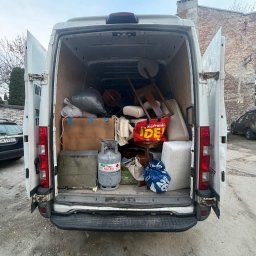 Clearbox pl - Wnętrze białego furgonetki wypełnionej różnymi przedmiotami, w tym meblami, workami i butlą gazową. Widok z tyłu, otwarte drzwi, tło miejskie z budynkiem z cegły.