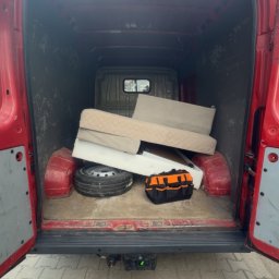 Clearbox pl - Czerwony van z otwartymi drzwiami bagażnika, wypełniony gruzem, oponą i torbą narzędziową. Widok z tyłu na kostkę brukową i fragment budynku w tle.