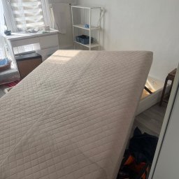 Clearbox pl - Materac z podnoszonym stelażem w jasnym pokoju, ukazujący schowek z narzędziami. Widoczne biurko, regał i krzesło. Neutralna kolorystyka, delikatne światło.