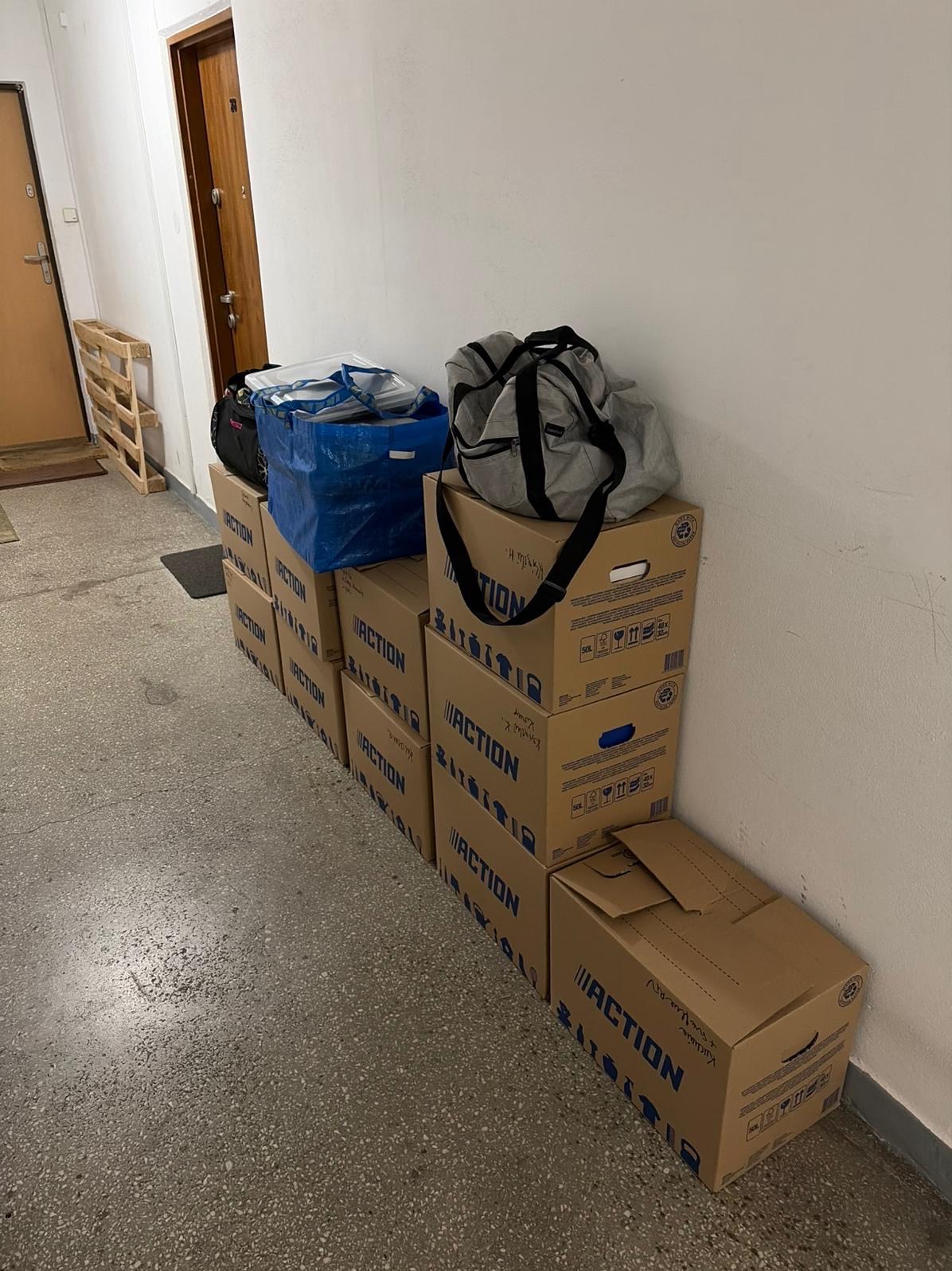 Stos kartonowych pudeł Action, torba podróżna i niebieska torba Ikea w korytarzu, prawdopodobnie podczas przeprowadzki. Widoczna paleta drewniana i drzwi.