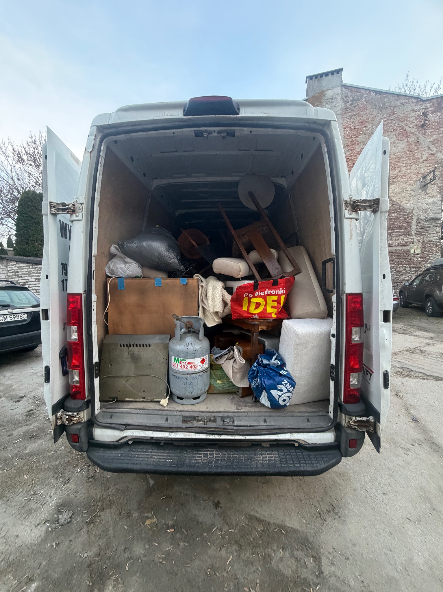 Wnętrze białego furgonetki wypełnionej różnymi przedmiotami, w tym meblami, workami i butlą gazową. Widok z tyłu, otwarte drzwi, tło miejskie z budynkiem z cegły.