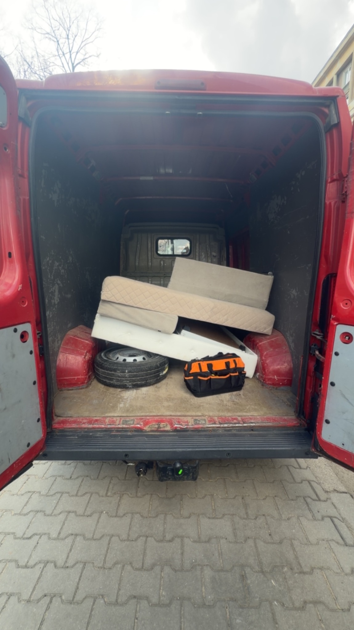 Czerwony van z otwartymi drzwiami bagażnika, wypełniony gruzem, oponą i torbą narzędziową. Widok z tyłu na kostkę brukową i fragment budynku w tle.