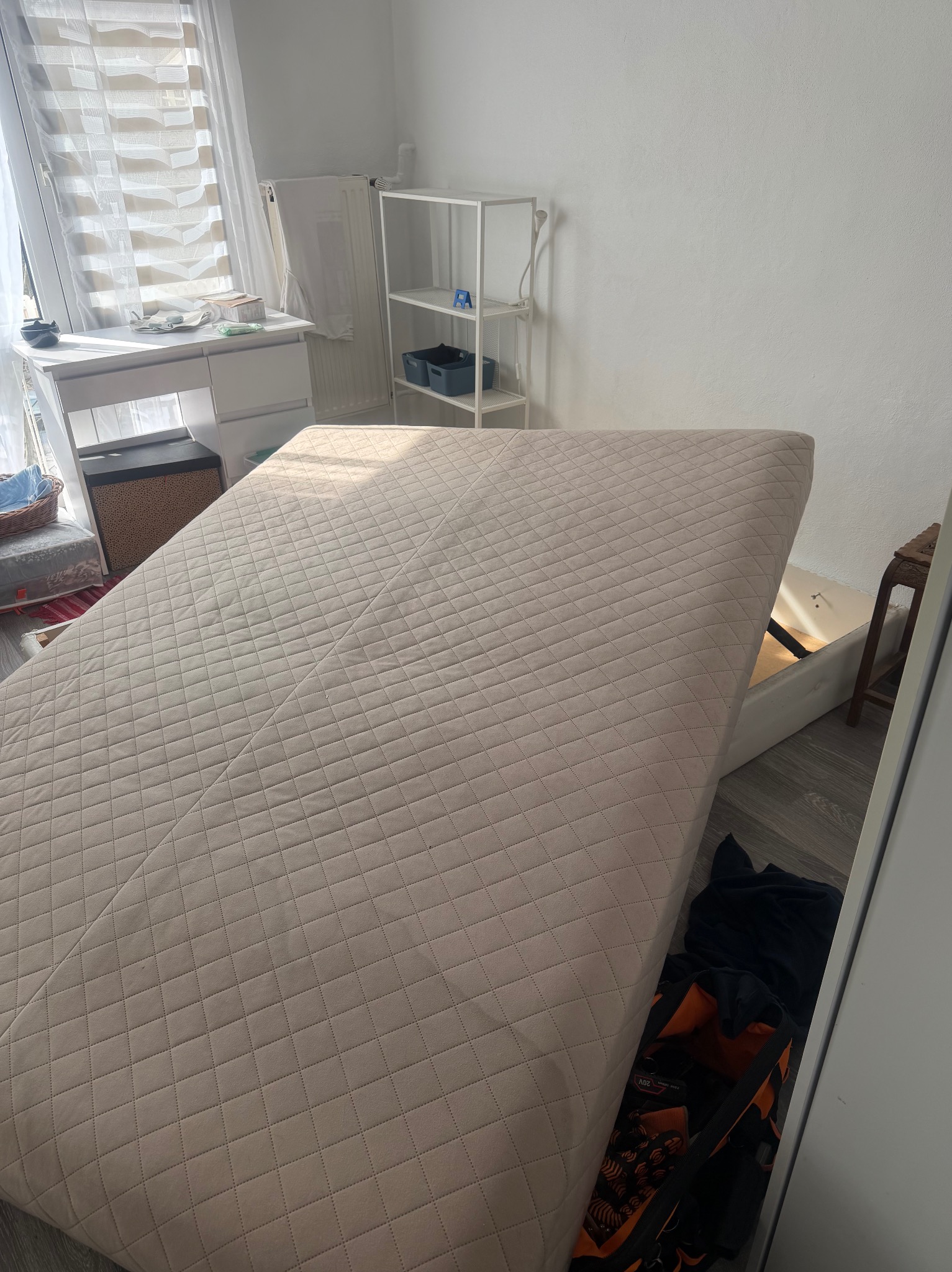 Materac z podnoszonym stelażem w jasnym pokoju, ukazujący schowek z narzędziami. Widoczne biurko, regał i krzesło. Neutralna kolorystyka, delikatne światło.