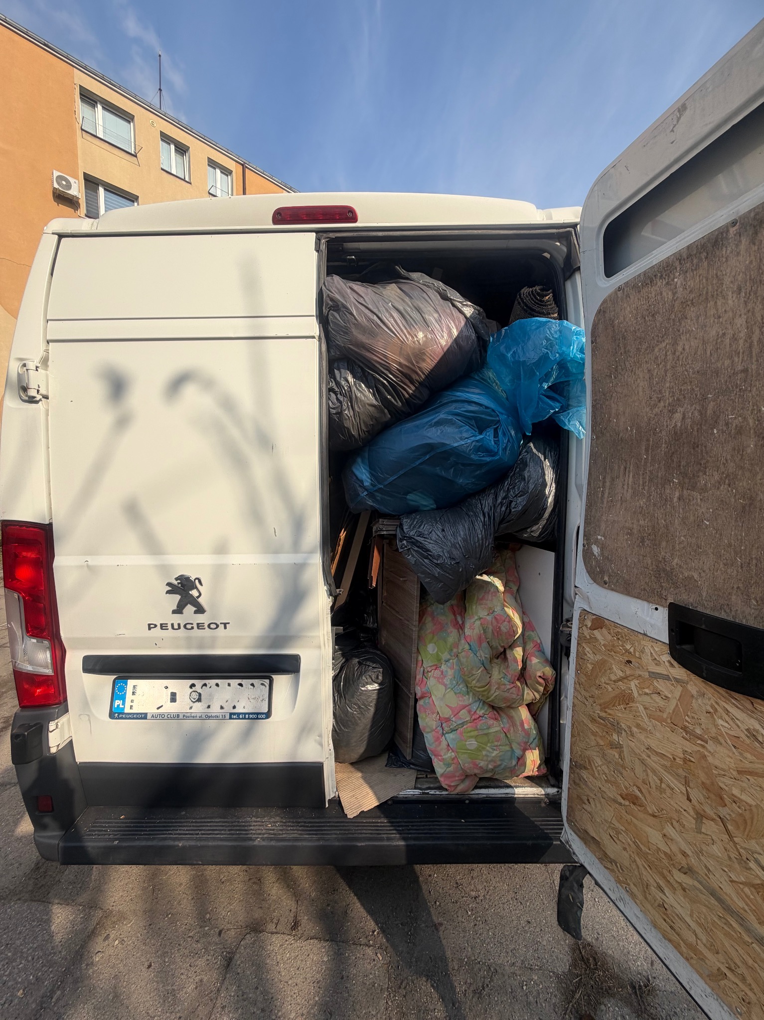 Peugeot wypełniony workami z odpadami po remoncie. Widoczne worki w kolorze czarnym i niebieskim, kołdra w kwiaty i elementy drewniane. Otwarty bagażnik.