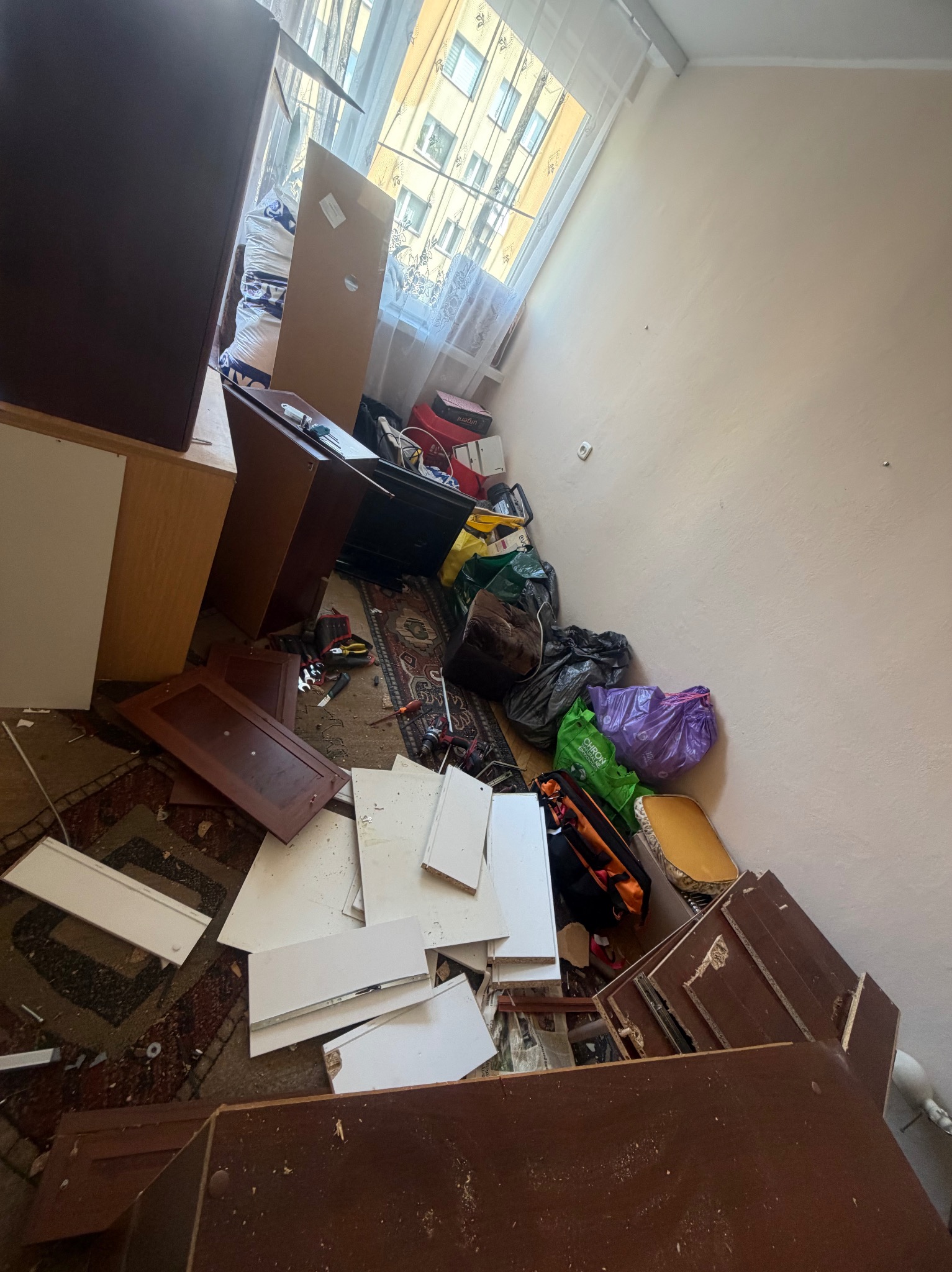 Pokój po demontażu mebli: porozrzucane elementy szafek, narzędzia, worki na śmieci, dywan. Widok z góry, ukośnie. Naturalne światło z okna.