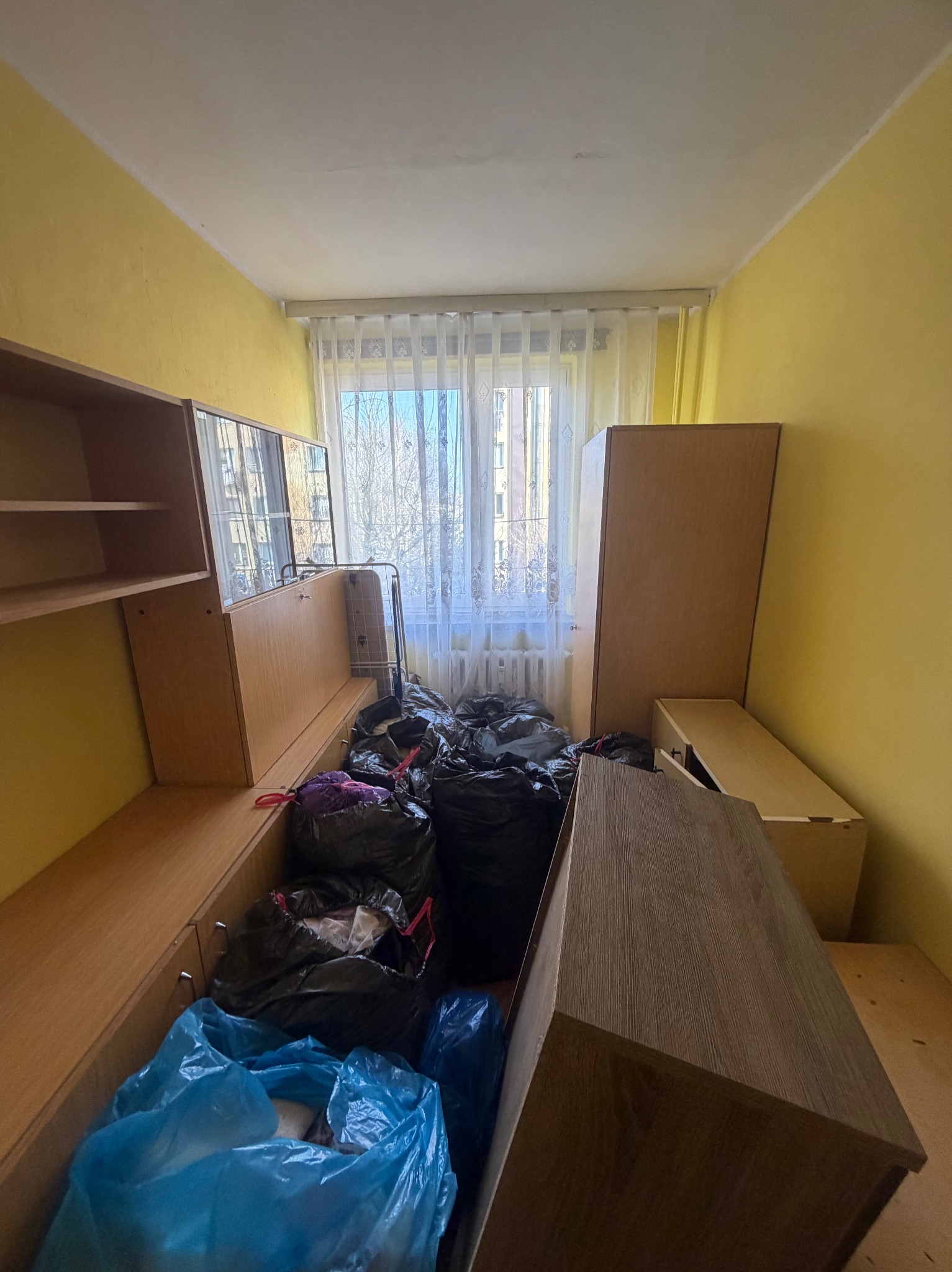 Pokój wypełniony workami ze śmieciami i meblami przed sprzątaniem. Widoczne żółte ściany i okno z firanką. Część mebli zdemontowana, przygotowana do wyniesienia.