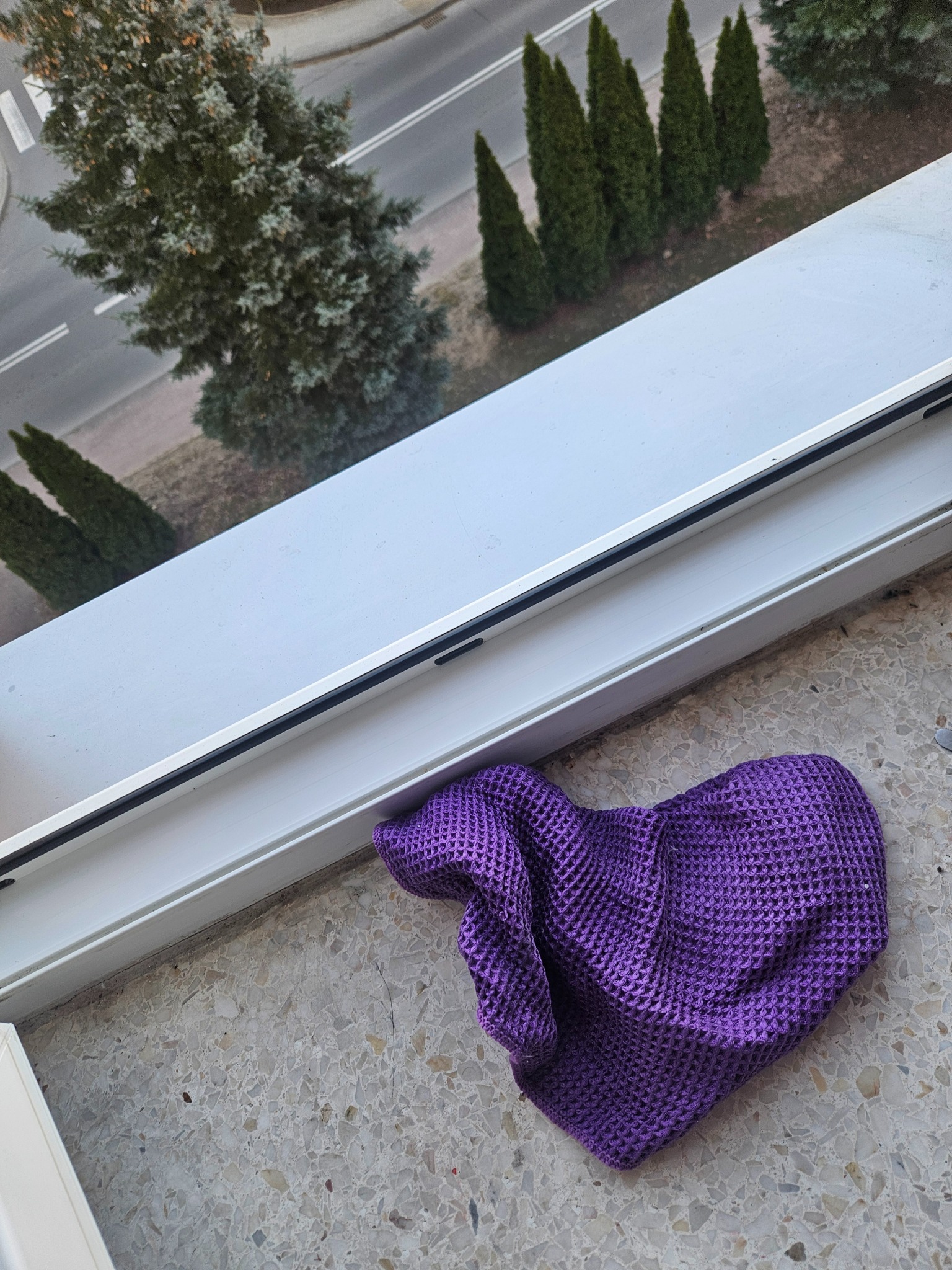 Fioletowa ściereczka leży na marmurowym parapecie okna z widokiem na drzewa i ulicę. Okno jest białe, a ściereczka ma strukturę wafla.