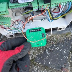 UNOVIA Michał Uczniak - Podłączenie modułu Wi-Fi SBW-02 do sterownika bramy. Widoczne złącza, przewody i ręka w roboczej rękawicy. Integracja inteligentnego sterowania bramą.