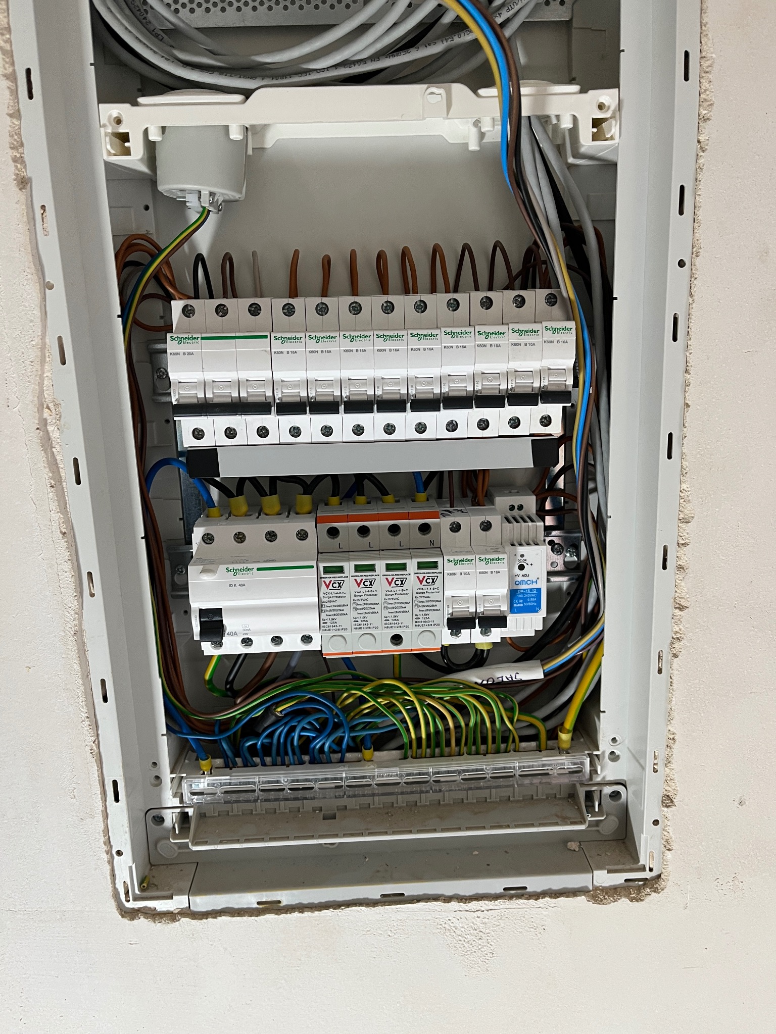 Rozdzielnica elektryczna z zabezpieczeniami przepięciowymi i wyłącznikami, widoczne przewody i elementy montażowe w białej obudowie.