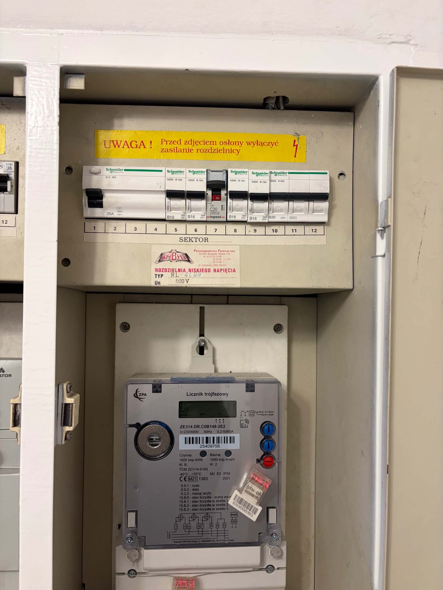 Otwarta skrzynka elektryczna z bezpiecznikami Schneider Electric, licznikiem trójfazowym ZPA i żółtą naklejką ostrzegawczą. Widoczne numery bezpieczników i oznaczenia napięcia.