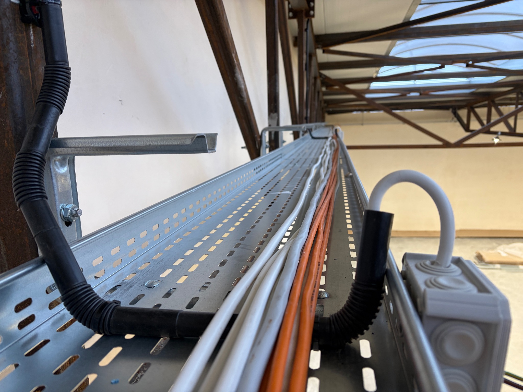 Metalowa korytka kablowa z ułożonymi przewodami elektrycznymi w kolorze białym i pomarańczowym, zamocowana do konstrukcji stalowej w hali.