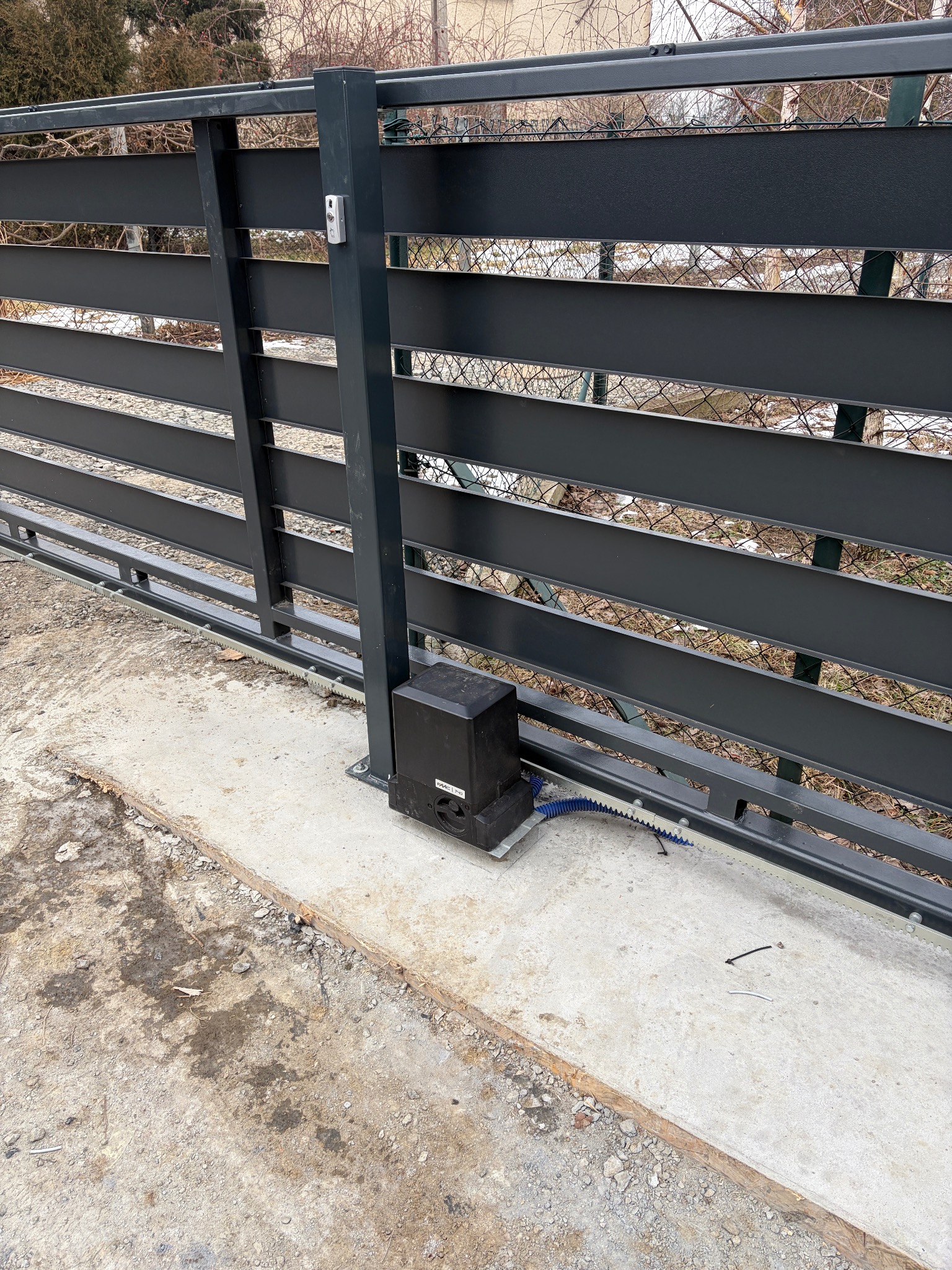 Nowoczesna, przesuwna brama w kolorze grafitowym z zamontowanym napędem i domofonem. Widoczna listwa zębata i przewody. Tło stanowią drzewa.