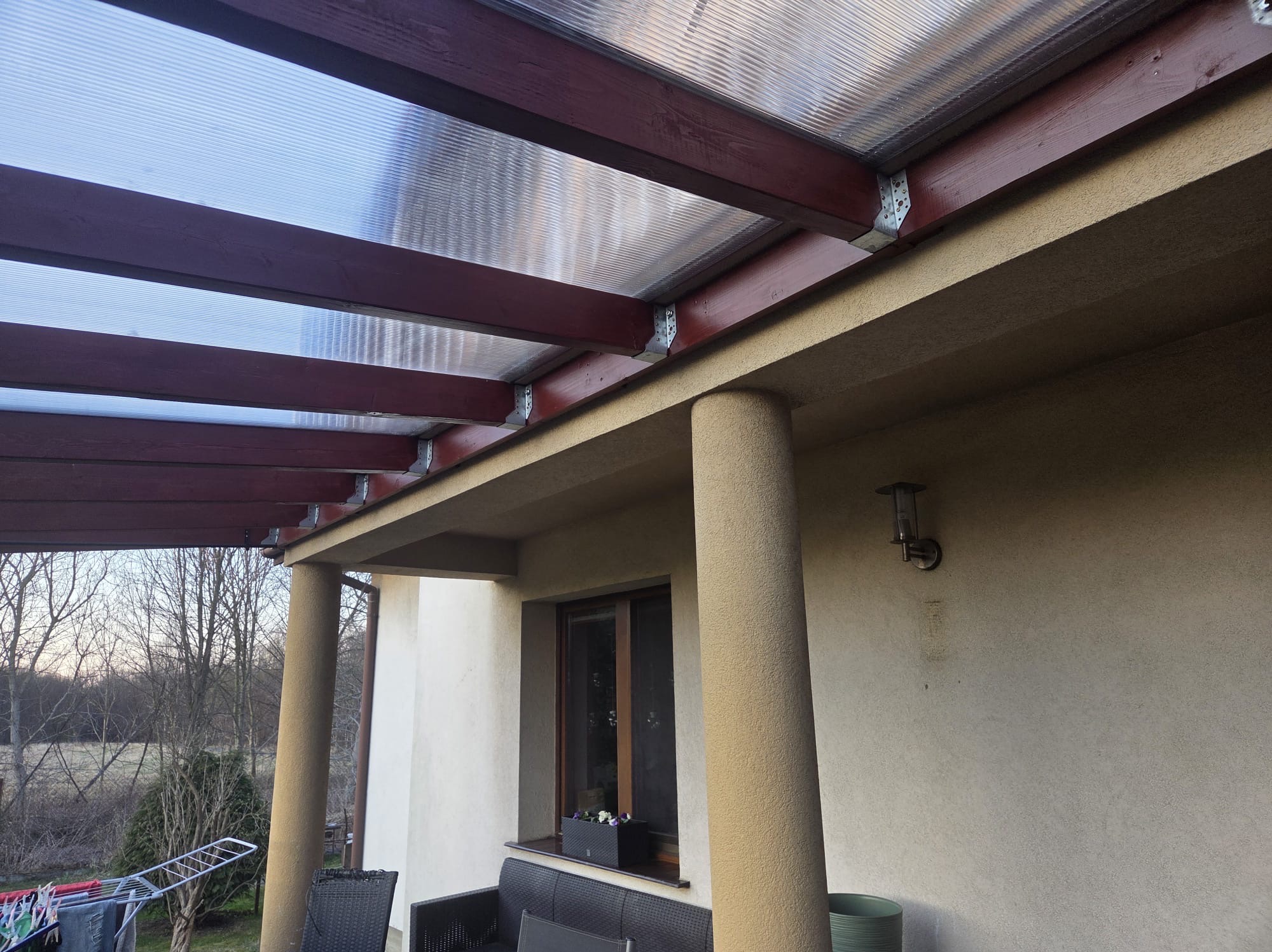 Zadaszenie tarasu z brązowymi belkami i przezroczystym poliwęglanem, wsparte na kolumnach. Widoczny fragment elewacji budynku i otoczenie.