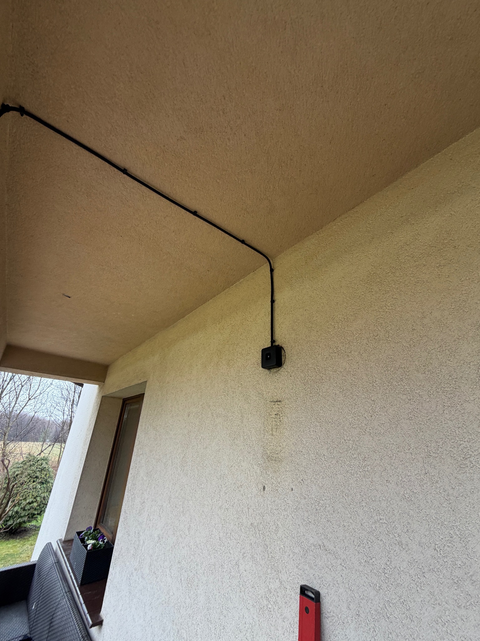 Czarna instalacja elektryczna natynkowa, poprowadzona wzdłuż beżowej ściany zewnętrznej budynku, z widoczną puszką przyłączeniową i fragmentem poziomicy.