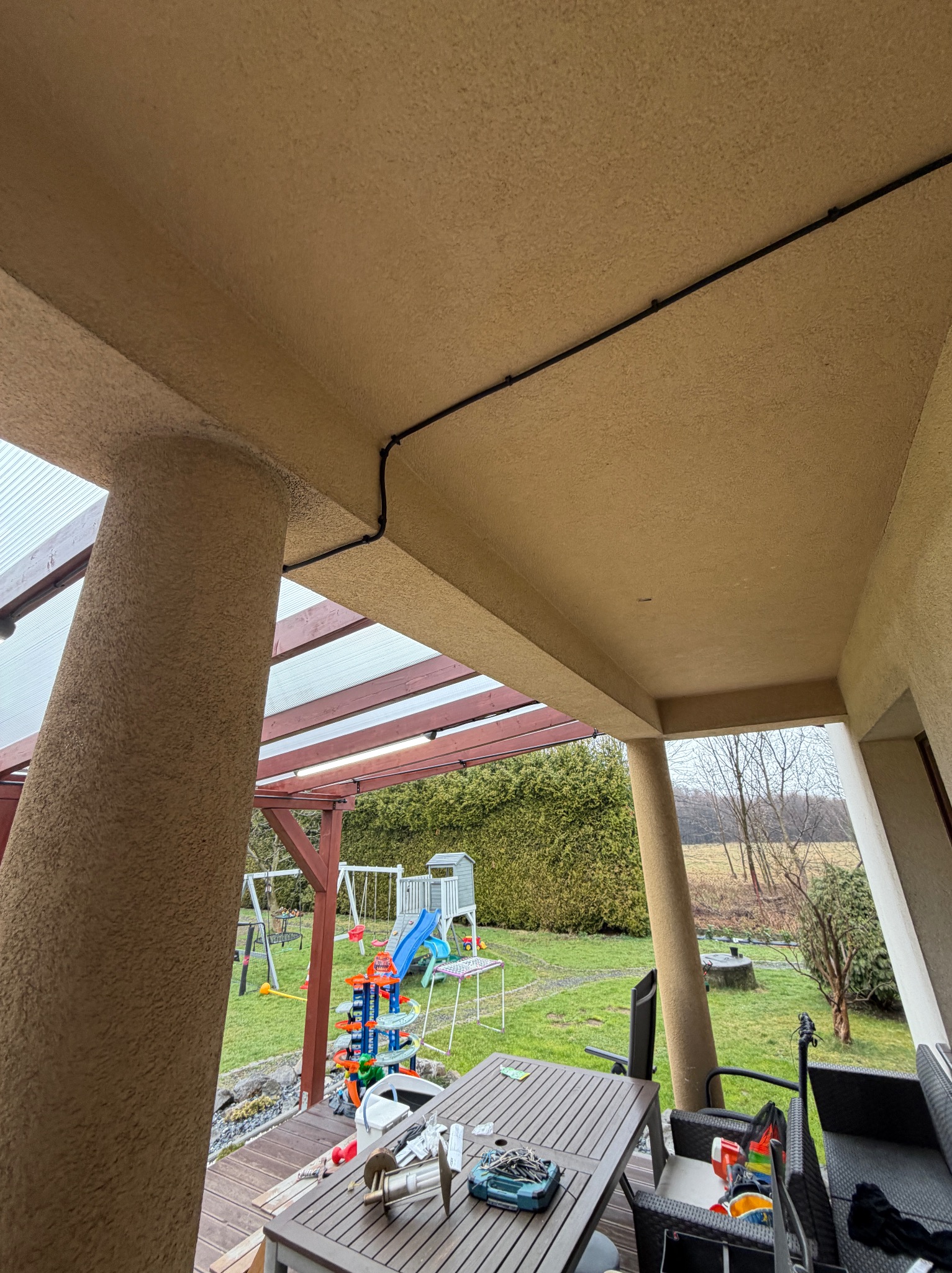 Czarna listwa z kablami przymocowana do beżowego sufitu z widocznymi elementami konstrukcyjnymi z drewna i ogrodem w tle. Widoczne narzędzia na stole.