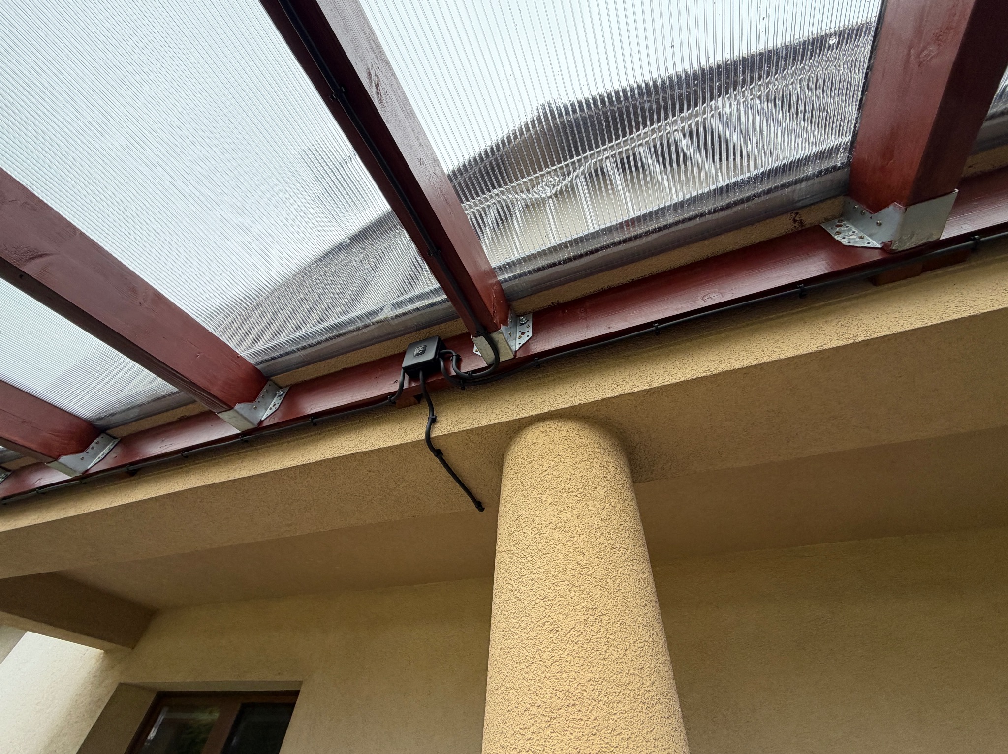 Zadaszenie tarasu z poliwęglanu, drewniana konstrukcja, widoczne mocowania i czarna puszka elektryczna z przewodami. Ujęcie od dołu, kolumna podtrzymująca.