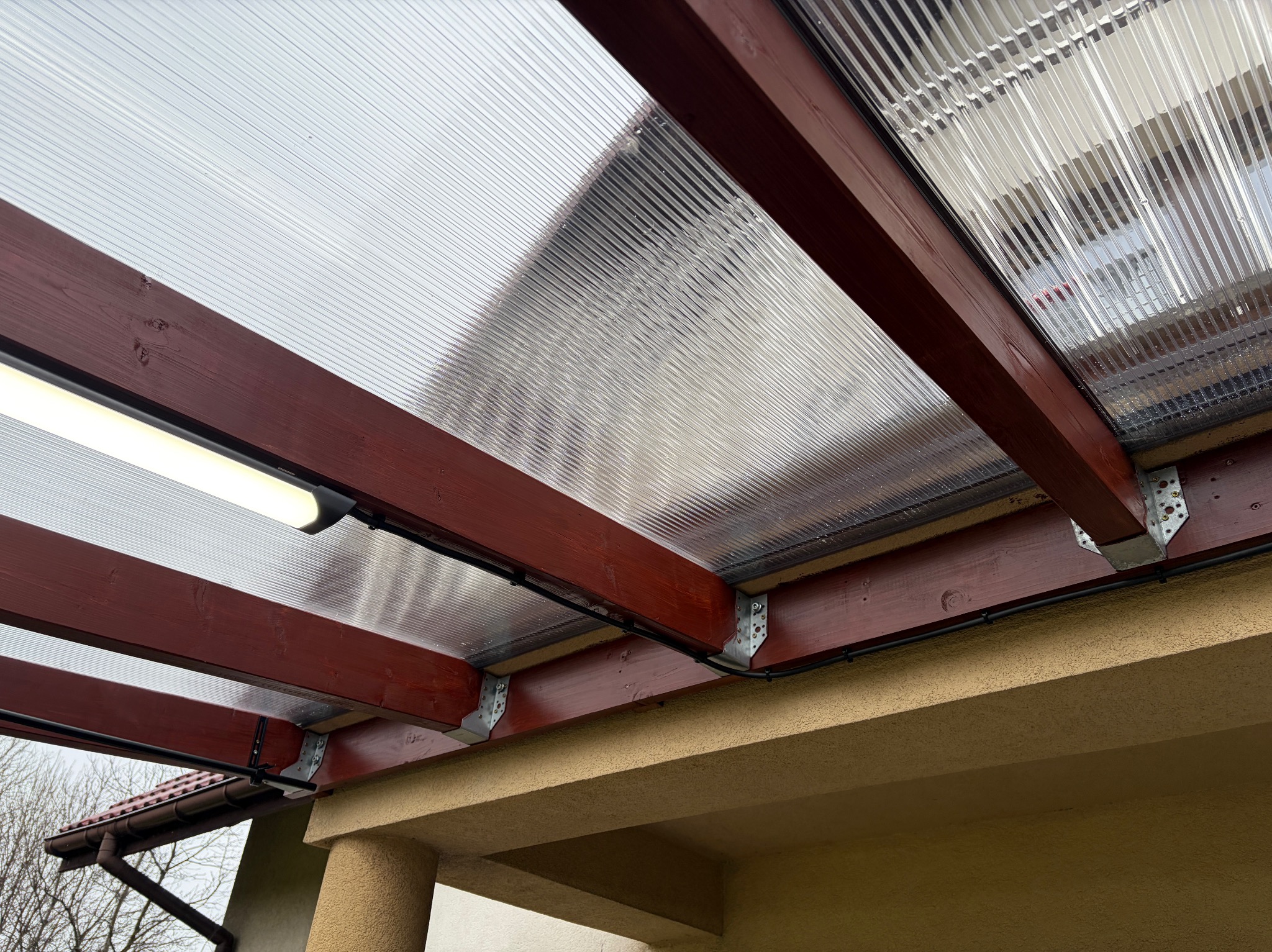 Zadaszenie tarasu z poliwęglanu, drewniana konstrukcja w kolorze mahoniu, oświetlenie LED. Widok z dołu, detale mocowań i instalacji elektrycznej.