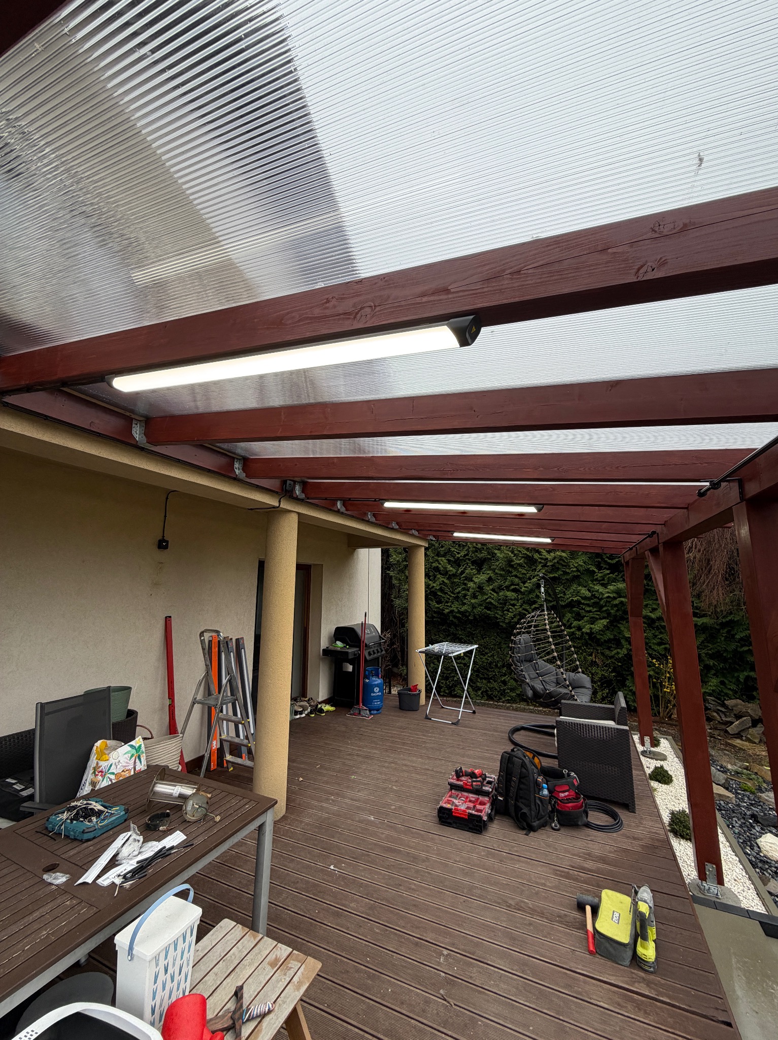 Zadaszenie tarasu z poliwęglanu z zamontowanym oświetleniem LED. Widoczne narzędzia i akcesoria na drewnianym tarasie. Ujęcie z dołu, szeroki kadr.