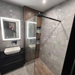 LUX-FREE - Nowoczesna łazienka z szarymi płytkami imitującymi marmur, kabiną prysznicową typu walk-in i podświetlanym lustrem. Widoczna umywalka nablatowa i drewniane akcenty.
