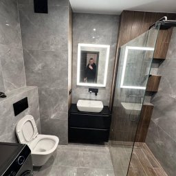 LUX-FREE - Nowoczesna łazienka z szarymi płytkami, podświetlanym lustrem i kabiną prysznicową typu walk-in. Umywalka nablatowa na czarnej szafce. Widoczne odbicie osoby w lustrze.